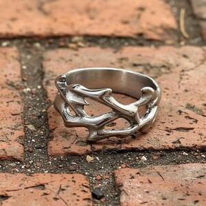 Peut inclure: Une bague argentée au design ouvert et organique. La bague présente un motif complexe et fluide avec une surface texturée. Le bijou est posé sur un fond de briques rouges, soulignant l'éclat métallique de la bague.