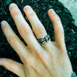 Può includere: Un anello argentato dal design geometrico è indossato su una mano. L'anello presenta una serie di forme rettangolari interconnesse. La mano è appoggiata su una superficie scura e strutturata, con un braccialetto d'argento visibile sul polso.
