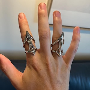 May include: Two silver-colored, articulated finger rings. Each ring features a pointed, triangular design with an open, web-like structure. The rings are worn on the index and middle fingers, showcasing a unique, edgy style.