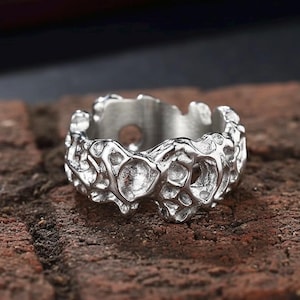 May include: A silver-colored ring with a textured, organic design. The ring features an irregular, porous surface, with a pattern of interconnected shapes. The ring is set against a brown brick surface.