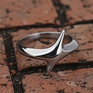 Puede incluir: Un anillo plateado pulido con un diseño abstracto único. El anillo presenta un elemento central puntiagudo que se extiende hacia arriba y hacia abajo, creando una forma distintiva. El anillo está sobre una superficie de ladrillo marrón rojizo texturizado.