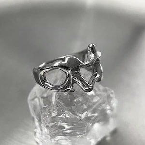 Peut inclure: Une bague argentée au design abstrait et fluide. La bague présente une forme ouverte et irrégulière avec plusieurs ouvertures. La bague est présentée sur une surface cristalline transparente, créant un contraste entre le métal lisse et la base texturée.