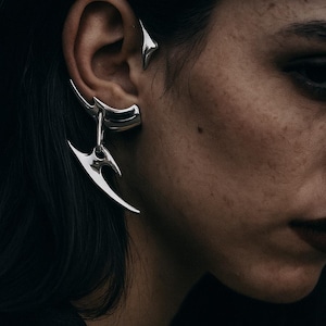 Puede incluir: Juego de aretes plateados. El conjunto incluye un brazalete de oreja curvado y un arete colgante con un diseño afilado y angular. Las joyas se usan en la oreja, con cabello oscuro y piel en el fondo.