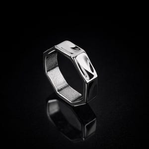 May include: A silver-colored ring with a geometric, hexagonal design. The ring has a polished, reflective surface and a textured interior. The ring is set against a black background, highlighting its shape and shine.