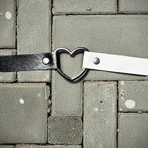 Puede incluir: Un collar gargantilla en blanco y negro con un centro en forma de corazón plateado. La correa negra está a la izquierda y la blanca a la derecha. Cada correa está asegurada con un perno plateado.