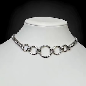 Puede incluir: Un collar gargantilla plateado con diseño de cadena y cinco colgantes circulares. El collar se exhibe sobre una cabeza de maniquí blanca sobre un fondo negro.