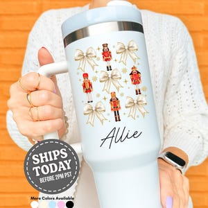 May include: White insulated tumbler featuring a festive design of nutcrackers and bows. The tumbler has a light blue lid and handle, with the name "Allie" in script. The tumbler is designed to keep beverages cold or hot.