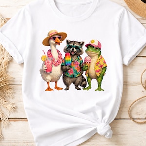 May include: White t-shirt featuring a whimsical illustration of a goose, raccoon, and frog, each wearing sunglasses and summer attire. The goose holds a pink drink, the raccoon a popsicle, and the frog an ice cream cone. The design evokes a fun, summery vibe.
