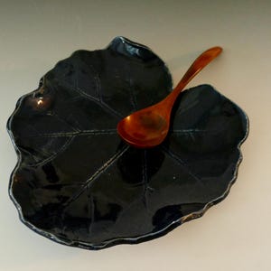 Op de afbeelding: Een donkerblauwe, blad-vormige keramische schaal met een glanzende afwerking. Een kleine houten lepel ligt op de schaal. De schaal heeft een textuur met zichtbare bladnerven en een licht verhoogde rand.