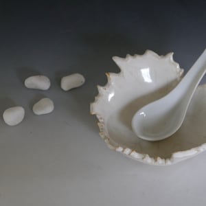 Puede incluir: Un cuenco blanco de cerámica con forma de hoja y una cuchara a juego. El cuenco tiene una superficie texturizada y un borde dorado. Cinco pequeñas piedras blancas lisas están dispersas cerca. Los artículos están dispuestos sobre un fondo degradado.