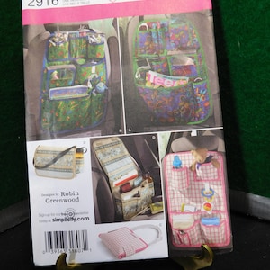May include: Simplicity sewing pattern number 2916 for a car seat organizer. The pattern includes instructions for three different styles: a green and blue floral print, a pink and white gingham print, and a beige and brown floral print. The pattern is for one size.