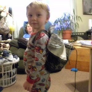 May include: A young boy wearing a grey and red pajama top with a camouflage backpack. The backpack has a long strap that is hanging down. The boy is standing in a room with a wooden table and a plant in a pot.