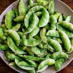 May include: A large, round, light-colored bowl filled with a heap of fresh, raw green hyacinth bean pods. The pods are curved, smooth, and vary in shades of light to medium green, showing natural textures and small stems at the ends.