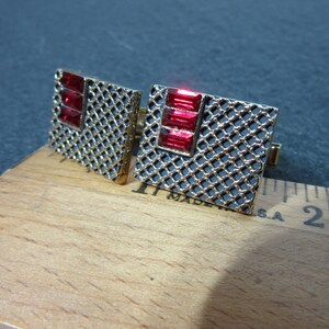 May include: A pair of gold cufflinks with a black mesh design and red rhinestone accents.