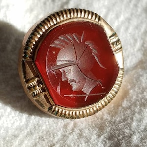 May include: A gold ring with a red carnelian stone featuring a carved image of a warrior wearing a helmet.