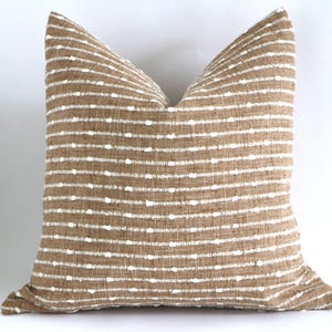 May include: A square decorative pillow with a tan and white striped pattern. The pillow has a textured fabric with horizontal tan stripes and evenly spaced white dots. The pillow is a neutral color and would complement any home decor.
