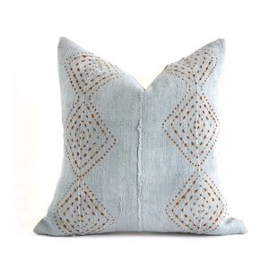May include: A square, pale blue cushion featuring a geometric diamond pattern in a warm brown hue. The cushion has a textured appearance, likely made from woven fabric. The design is symmetrical, with the diamond pattern repeated on both sides.