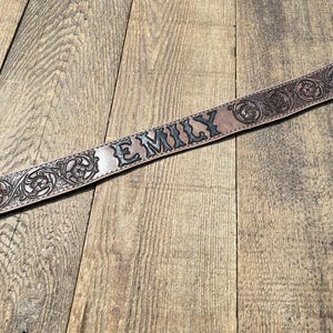 May include: A brown leather belt with the name "EMILY" embossed in black lettering. The belt features intricate floral and scrollwork designs along its edges, showcasing a detailed, handcrafted aesthetic.