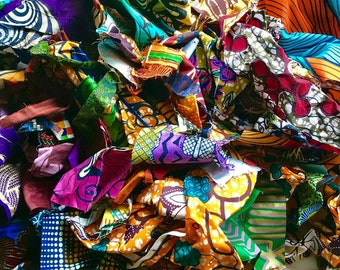 May include: A pile of colorful fabric scraps in various patterns and colors, including purple, orange, green, and yellow. The fabric pieces are cut into irregular shapes and sizes, creating a textured and layered appearance. The image showcases a variety of African wax print textiles.