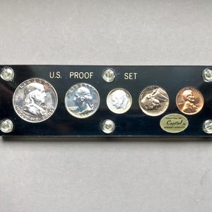 May include: A U.S. Proof Set displayed on a black rectangular holder. The set includes five different coins: a silver half dollar, a quarter, a dime, a nickel, and a penny. The holder is labeled "U.S. PROOF SET" and includes a "Capital" label.