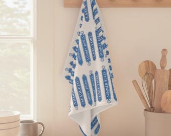 May include: A white kitchen towel featuring a repeating pattern of blue rectangular signs with rounded ends. Each sign contains text including "LOONEY DUNES," "LAZY FAIRE," "SUMMER RULES," "SHORE THING," "SEADUCED," and "VITAMIN SEA" in blue.