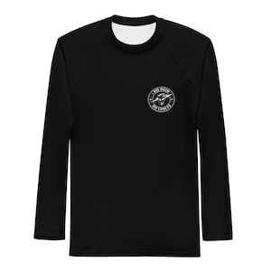 Può includere: Maglia sportiva nera a maniche lunghe con girocollo bianco e maniche a raglan. Un logo circolare bianco sul petto recita "NO PAIN NO LIMITS" attorno a un pugno chiuso. La maglia è progettata per attività sportive.