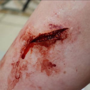 Può includere: Un'applicazione di trucco protesico realistica che raffigura una lacerazione profonda e aperta su pelle pallida. La ferita presenta bordi frastagliati e strutturati con sangue rosso scuro dall'aspetto umido all'interno dell'incisione.