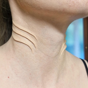 May include: A beige prosthetic appliance applied to the neck, featuring three raised, parallel, curved ridges that mimic the appearance of gills or layered skin folds, blending into the natural skin tone of the neck area.