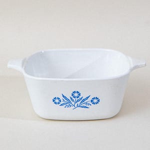 May include: White, square casserole dish with two handles. The dish features a blue floral design on the front. A vintage kitchenware item, perfect for baking and serving.