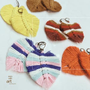 May include: A collection of handmade macrame feather earrings in various colors. The earrings are in shades of yellow, brown, red, white, blue, pink, and orange. Each earring has a metal hook for easy wear. The earrings are displayed on a white surface.