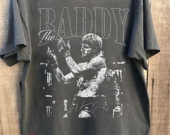May include: A charcoal grey short-sleeve t-shirt featuring a white graphic print of a fighter in a ring. The text "The BADDY" is printed in large white letters above the figure. The shirt has a crew neckline and a standard fit design.