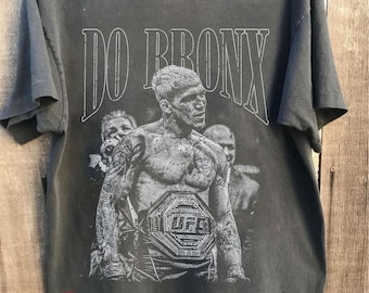 May include: Dark gray t-shirt featuring a black and white graphic of a UFC fighter with a championship belt. The text "DO BRONX" is above the image. The shirt has a "Made in USA" sticker and "COMFORT COLORS PEPPER" text.