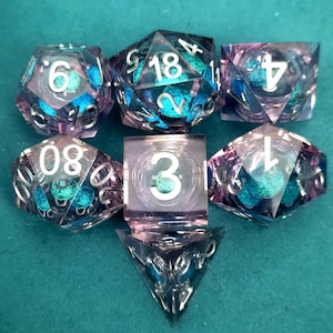 May include: A set of seven polyhedral dice with a transparent, iridescent design. The dice feature a mix of purple, blue, and silver colors, with white numbers. The set includes a D4, D6, D8, D10, D12, D20, and a cube.