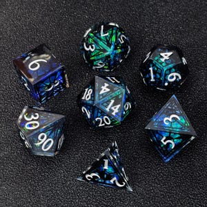 May include: A set of seven polyhedral dice featuring a black geometric cage overlay and a translucent blue and green iridescent core. Each die has white engraved numbers, including a d20, d12, d10, d8, d6, d4, and a percentile die.