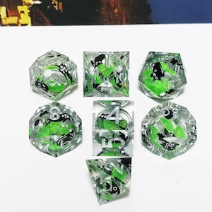 May include: Set of seven clear resin dice with green and black accents. Each die features a unique shape and number, with a green core and black octopus-like designs. Ideal for tabletop gaming.
