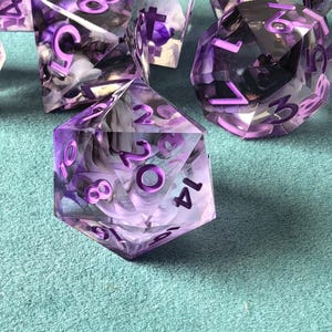 May include: Close-up of several polyhedral dice with a purple and white marbled design. The dice have white numbers and are set against a teal background. The dice are used for tabletop games.