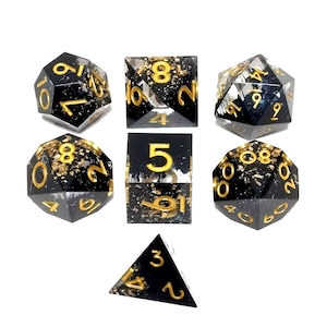 May include: Set of seven polyhedral dice in various shapes. The dice are black with gold-colored numbers and gold leaf inclusions. The dice are arranged on a white background.