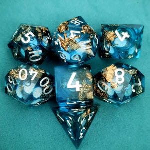 May include: A set of seven polyhedral dice in various shapes. The dice are a translucent blue with gold leaf inclusions and white numbering. The dice are arranged on a teal surface, showcasing their unique design and craftsmanship.