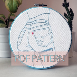 May include: Embroidered hoop art featuring a close-up of a pair of blue jeans with a red back pocket. The embroidery is done in blue thread on white fabric.