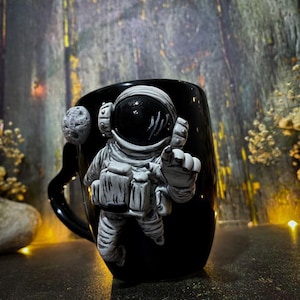 May include: Black ceramic mug featuring a white and grey astronaut figure and a small grey moon. The astronaut is in a spacesuit, with a raised hand. The mug has a black handle.