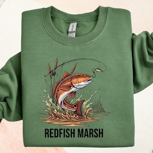 May include: Green sweatshirt featuring a graphic of a redfish jumping out of water, with a fishing rod and lure. The text "REDFISH MARSH" is printed below the image. The sweatshirt is a solid green color.