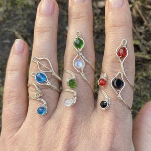 以下が含まれることがあります： 手には、手作りのワイヤーラップリングがいくつかあります。リングには、青、緑、赤、黒など、さまざまな色の宝石が使用されています。ワイヤーはシルバー色で、宝石は丸型または涙型です。