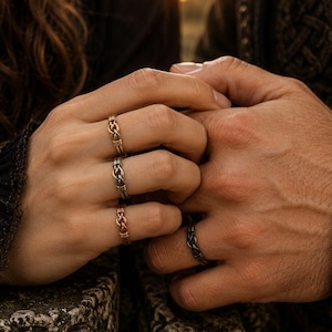 Puede incluir: Tres anillos metálicos con intrincados patrones trenzados en los dedos de una mano, y un anillo metálico oscuro con una textura trenzada similar en un dedo de la otra mano, ambas manos unidas en un abrazo cercano y entrelazado.