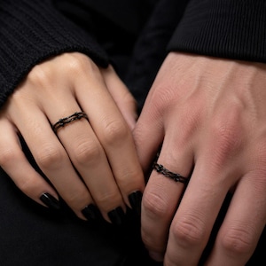 May include: A pair of thin, black metal rings featuring a textured design that mimics the appearance of barbed wire. The rings are worn on the fingers, showcasing a dark, metallic finish with a continuous, twisted wire-like pattern around the band.