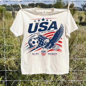 May include: A cream-colored short-sleeve t-shirt featuring a graphic of a bald eagle, a soccer ball, and the American flag. The text reads USA, ONE NATION ONE TEAM ONE DREAM, Est. 1913, and World Game '26, with a small shield logo at the bottom.