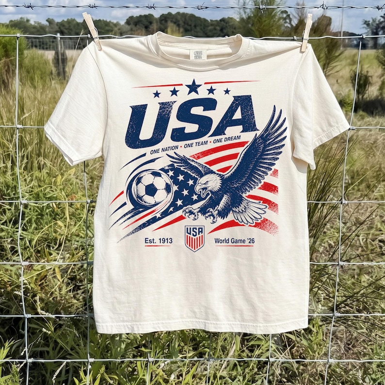 May include: A cream-colored short-sleeve t-shirt featuring a graphic of a bald eagle, a soccer ball, and the American flag. The text reads USA, ONE NATION ONE TEAM ONE DREAM, Est. 1913, and World Game '26, with a small shield logo at the bottom.