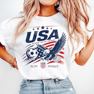 May include: A white t-shirt featuring a graphic print with the text USA, ONE NATION ONE TEAM ONE DREAM, Est. 1913, and World Game '26. The design includes a bald eagle, a soccer ball, and a stylized American flag with red and blue elements.