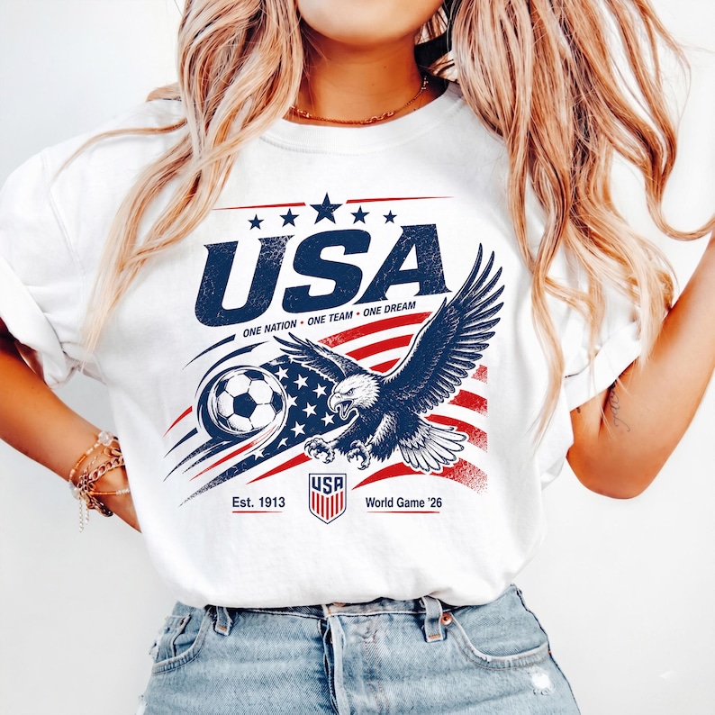 May include: A white t-shirt featuring a graphic print with the text USA, ONE NATION ONE TEAM ONE DREAM, Est. 1913, and World Game '26. The design includes a bald eagle, a soccer ball, and a stylized American flag with red and blue elements.
