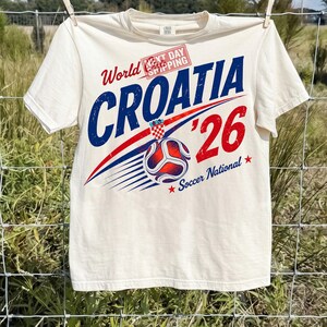 May include: A cream-colored short-sleeve t-shirt featuring a graphic design with the text World Croatia '26 Soccer National. The design includes a soccer ball, a red and white checkered emblem, and a red stamp reading Next Day Shipping.