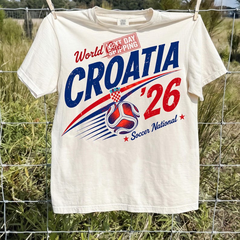 May include: A cream-colored short-sleeve t-shirt featuring a graphic design with the text World Croatia '26 Soccer National. The design includes a soccer ball, a red and white checkered emblem, and a red stamp reading Next Day Shipping.